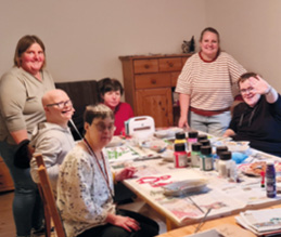 Eine Gruppe Menschen mit Behinderungen sitzen am Tisch und malen. 2 Betreuer stehen dabei.