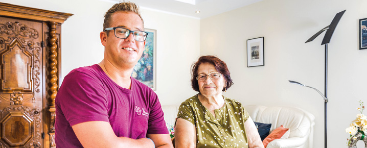 Ein Pfleger und eine Senorion sitzen nebeneinander und schauen in die Kamera.